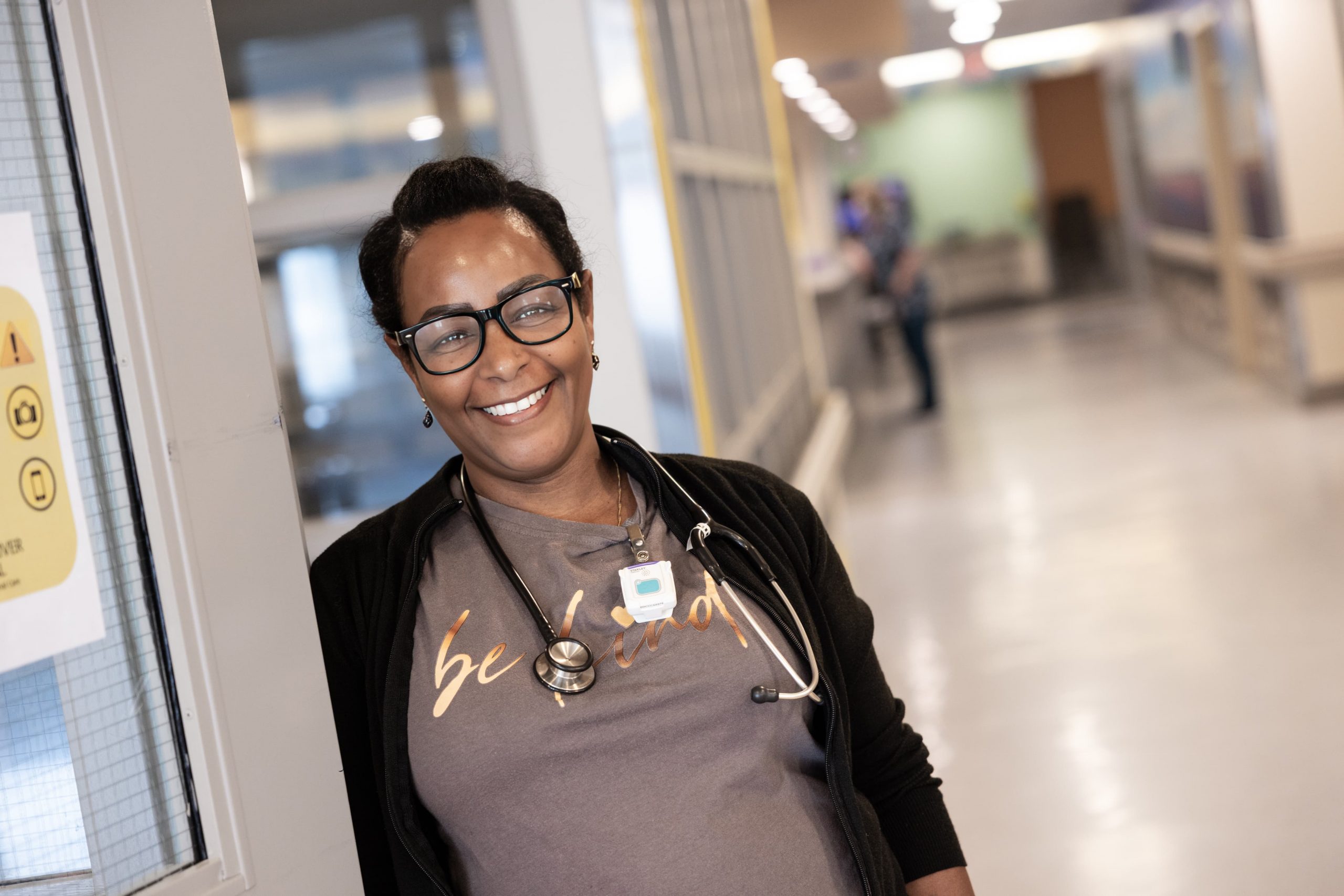 A smiling healthcare worker wearing a stethoscope stands in a hospital hallway near a door.
