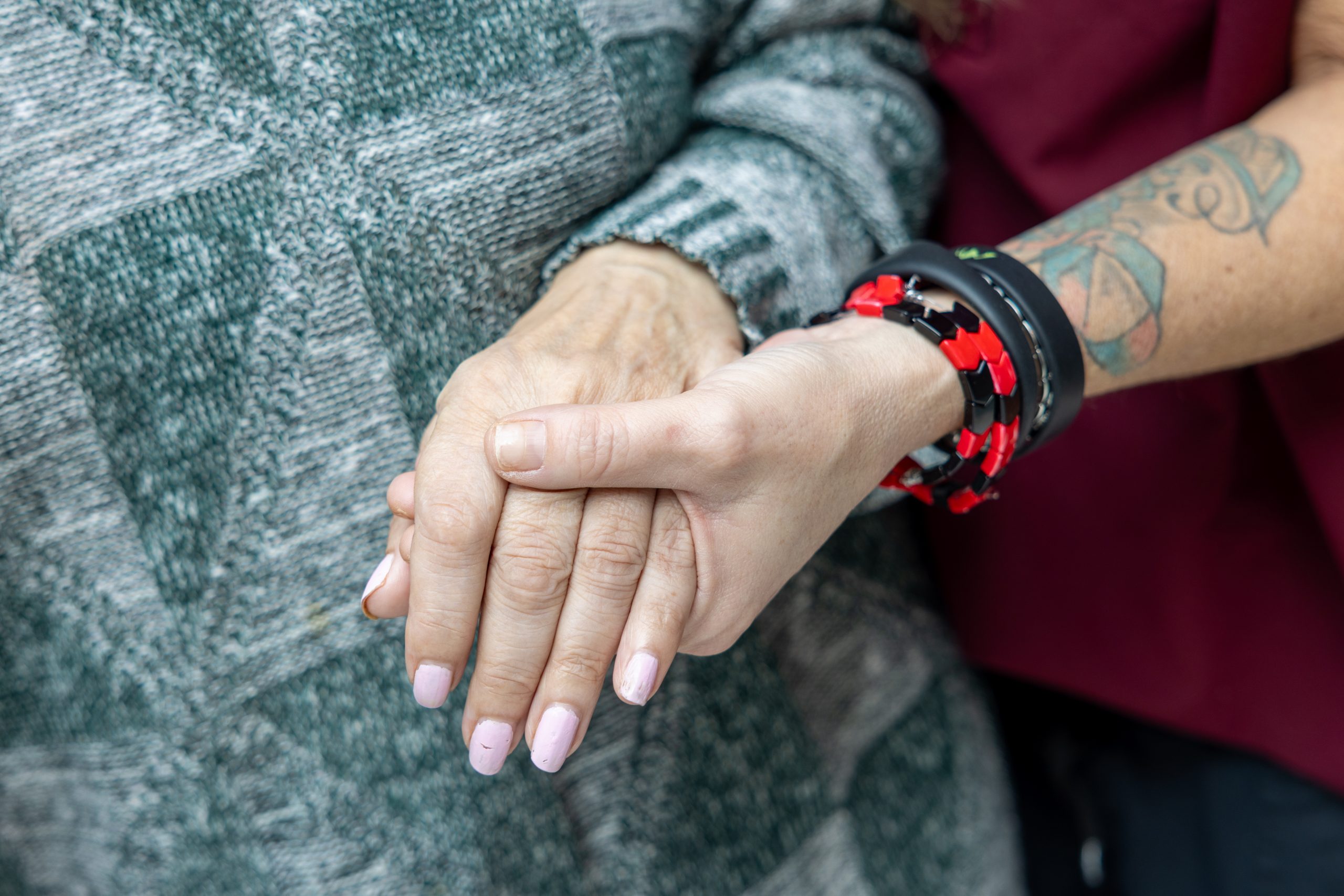 Two people hold hands; one wears a gray sweater, the other has a tattoo and wears a red and black bracelet.