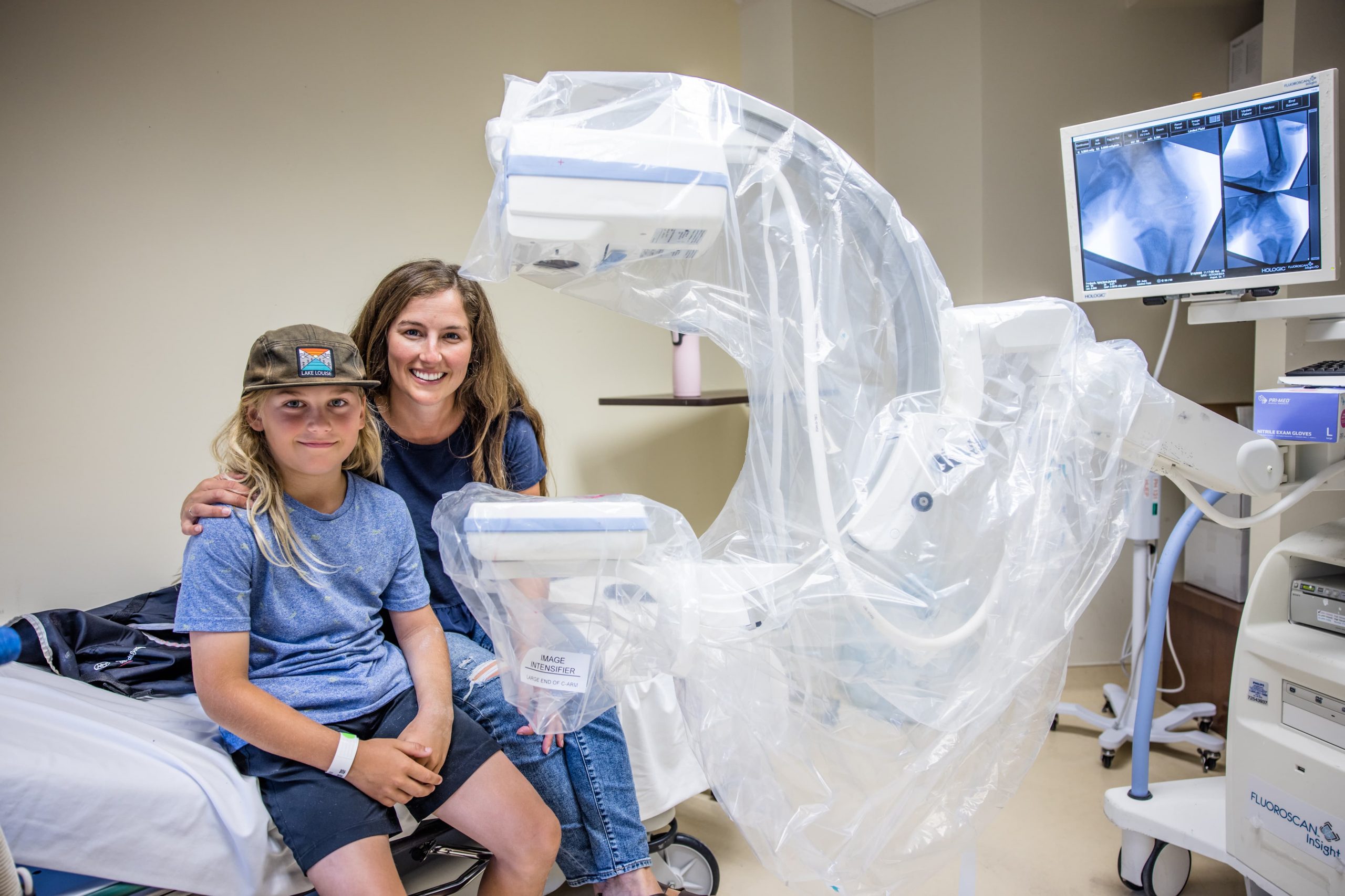 A woman and a child sit next to a medical imaging machine in a hospital room with X-ray images on a monitor.