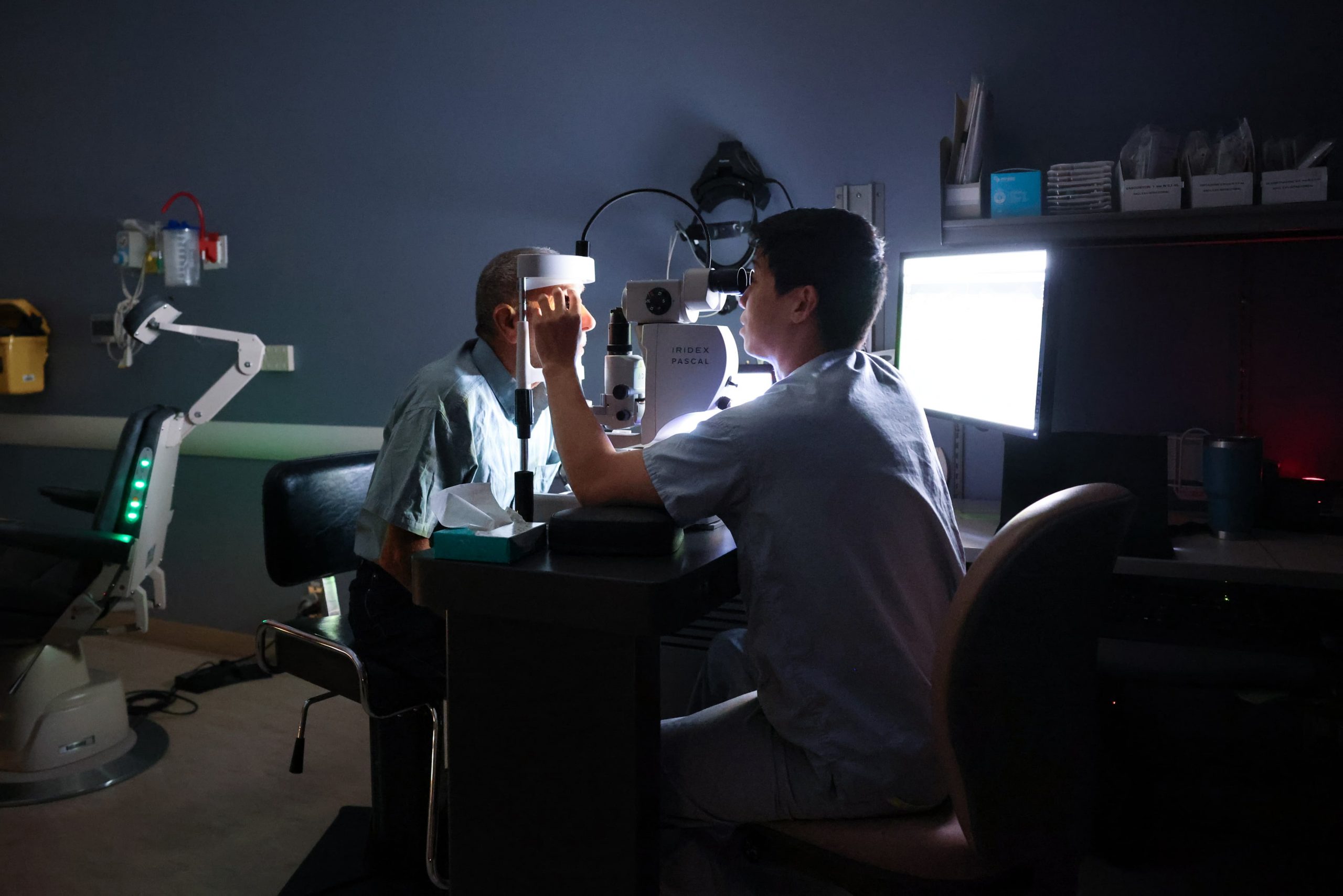 An eye doctor examines a patient's eyes using a slit lamp in a dimly lit clinic room.