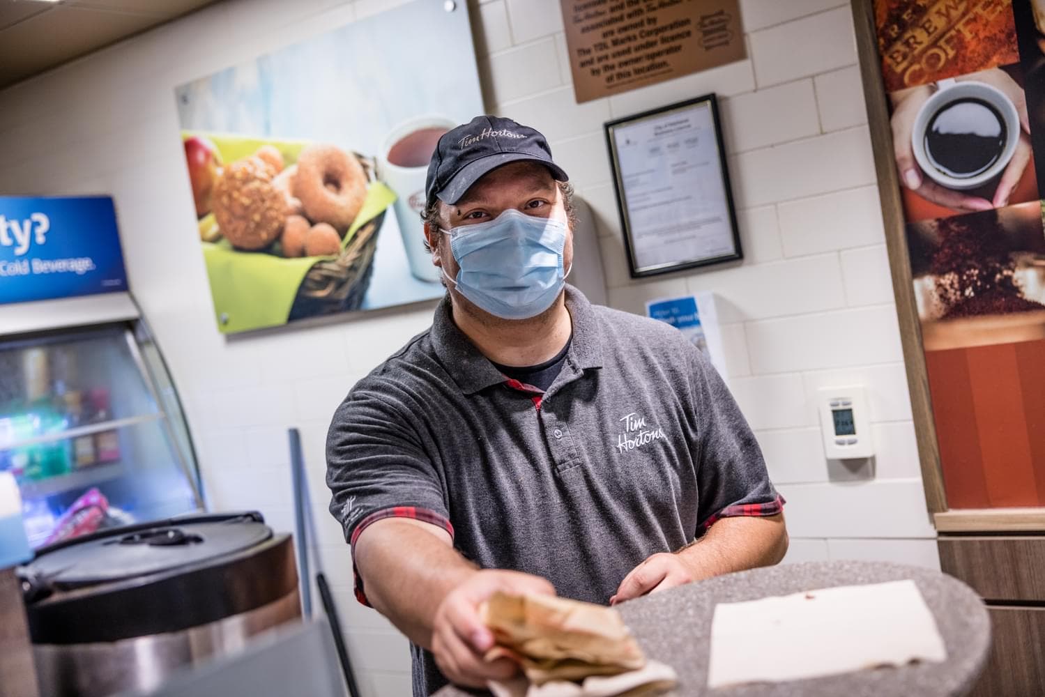 A Tim Hortons employee wearing a mask hands a paper bag to a customer at the counter.