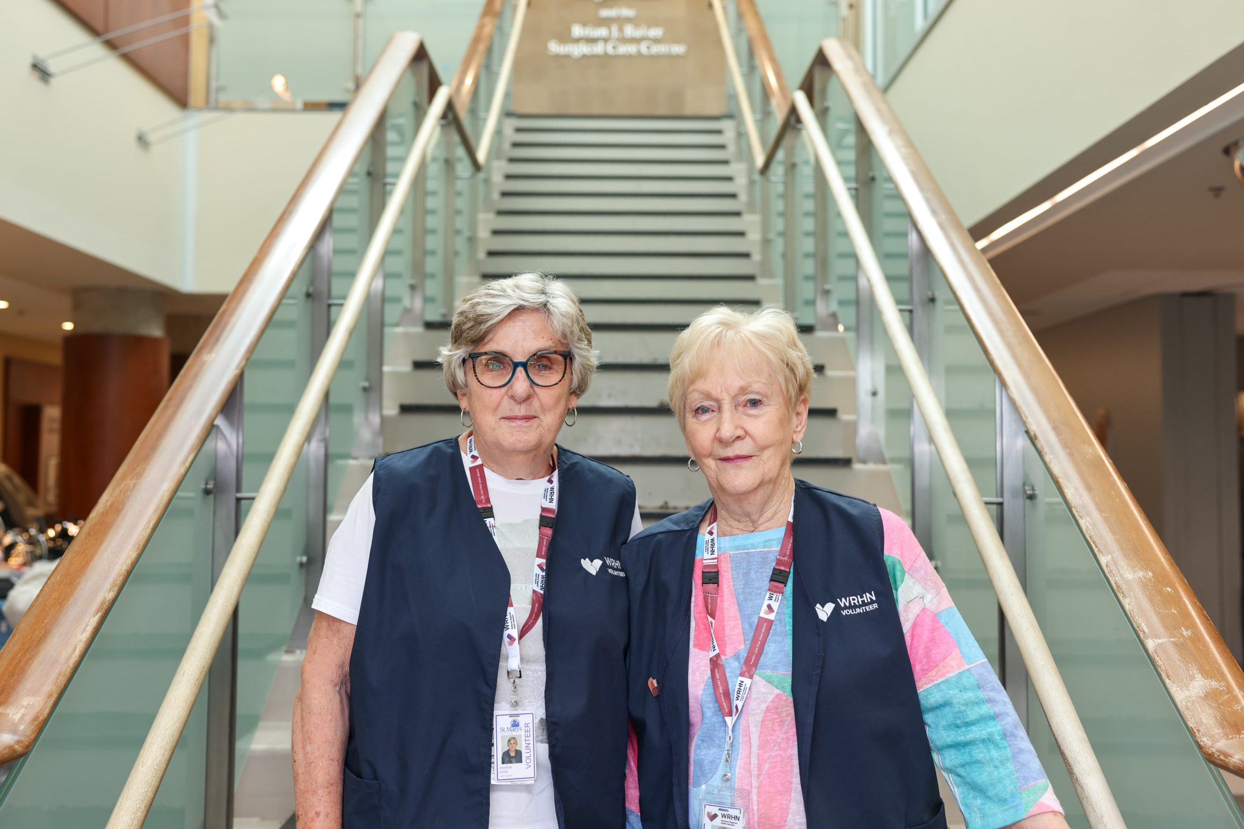 Two women wearing volunteer vests and name badges stand in front of a staircase inside a building.