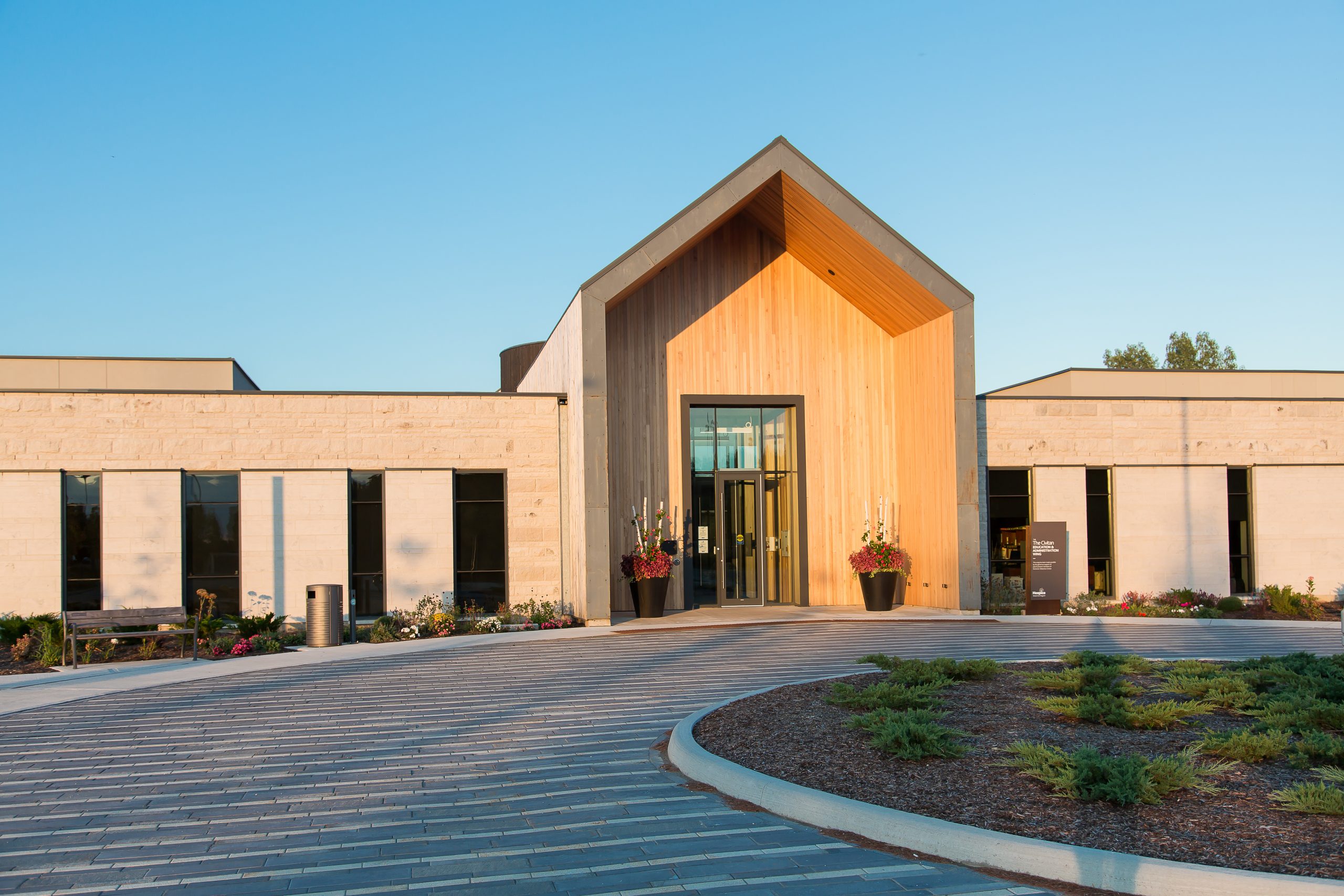 Modern building with stone and wood exterior, large glass entrance, and landscaped walkway under clear sky.