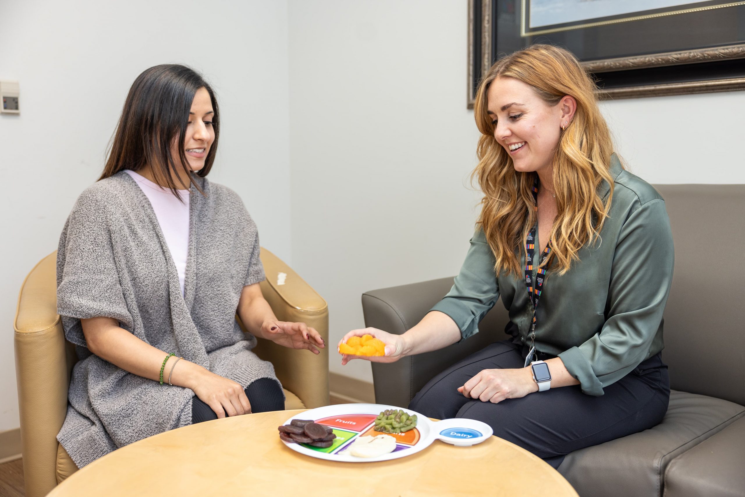 Two women sit and discuss food portion sizes using a divided plate and food models on a small table.