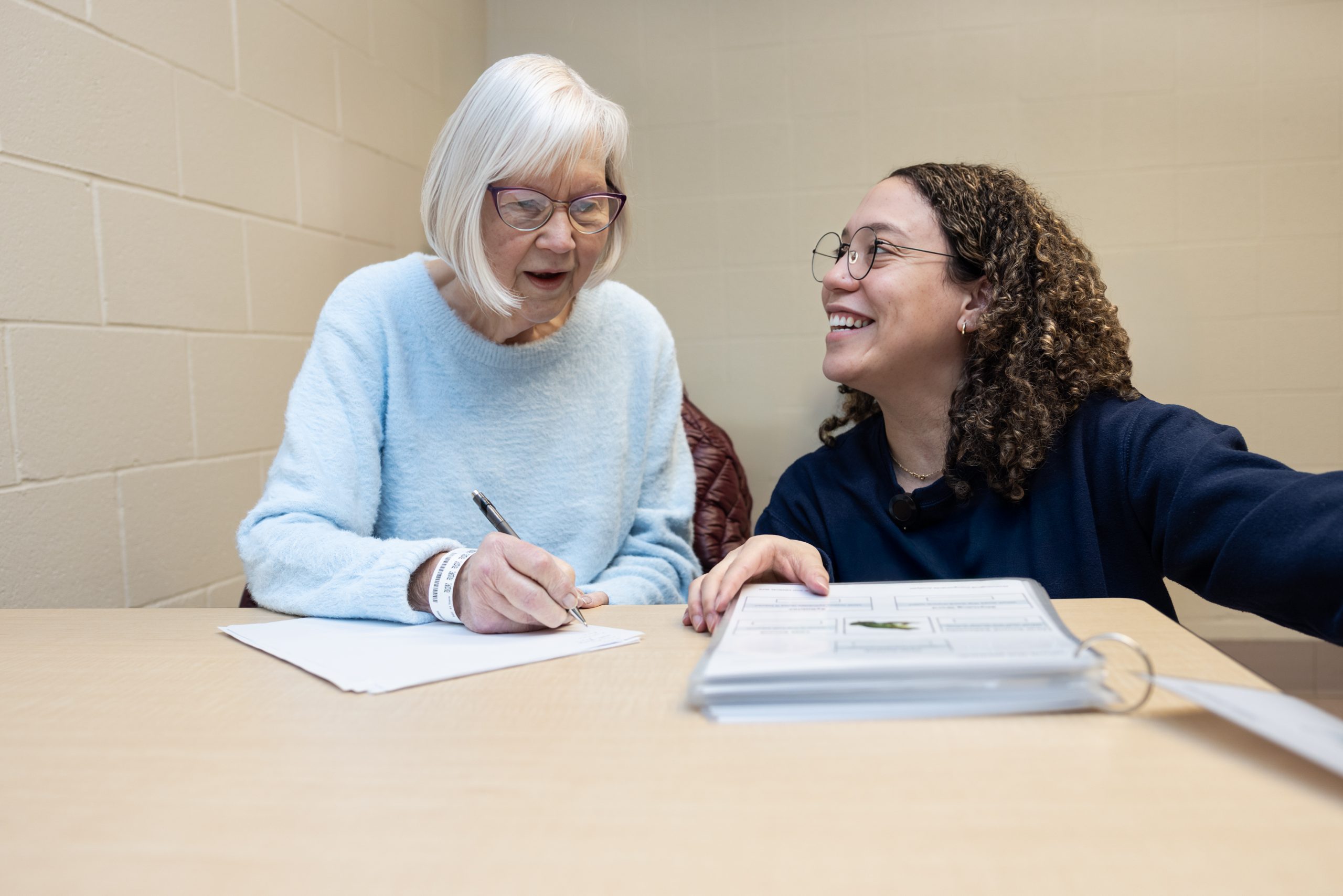 An older woman writes on paper while a younger woman smiles and holds a binder at a table in a plain room.