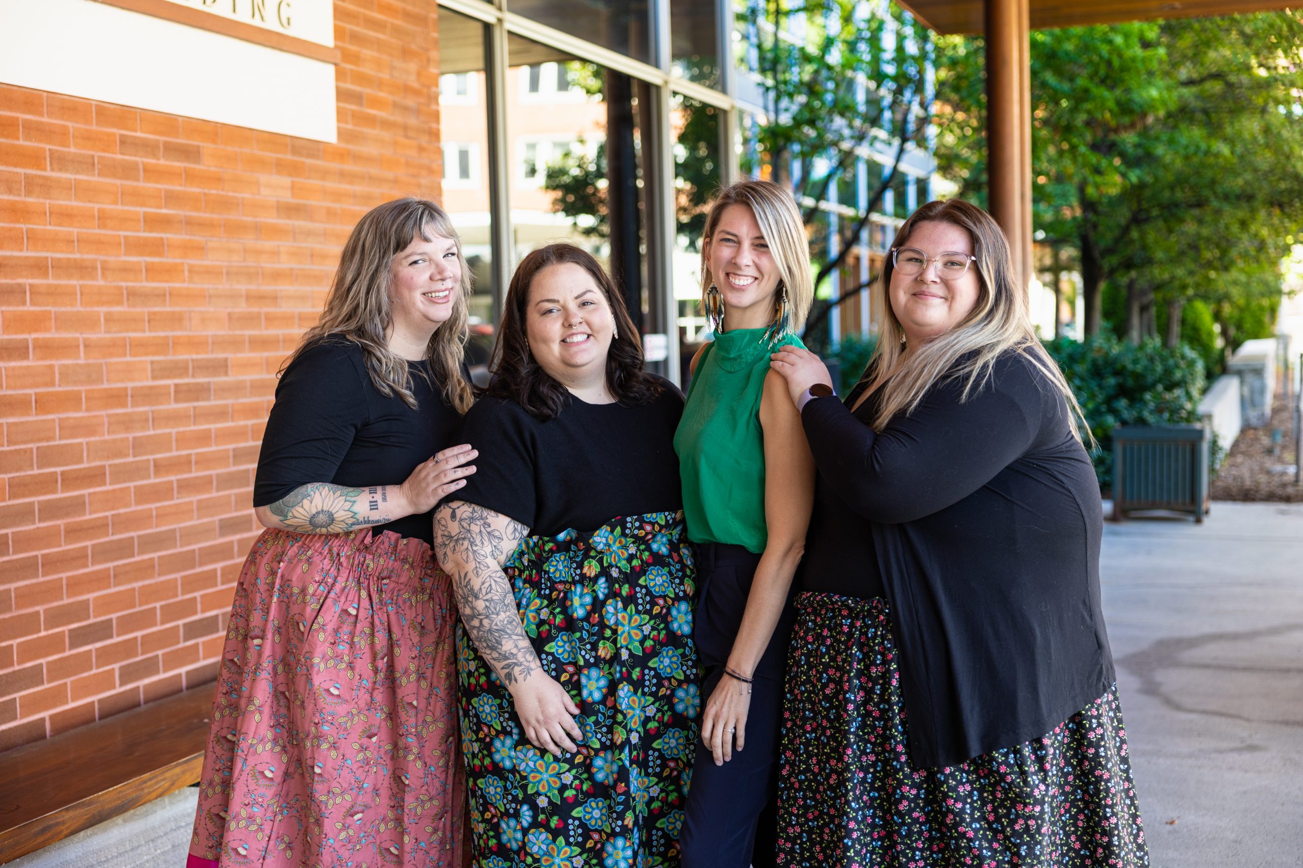 Four women stand together outside a brick building, smiling at the camera on a sunny day.
