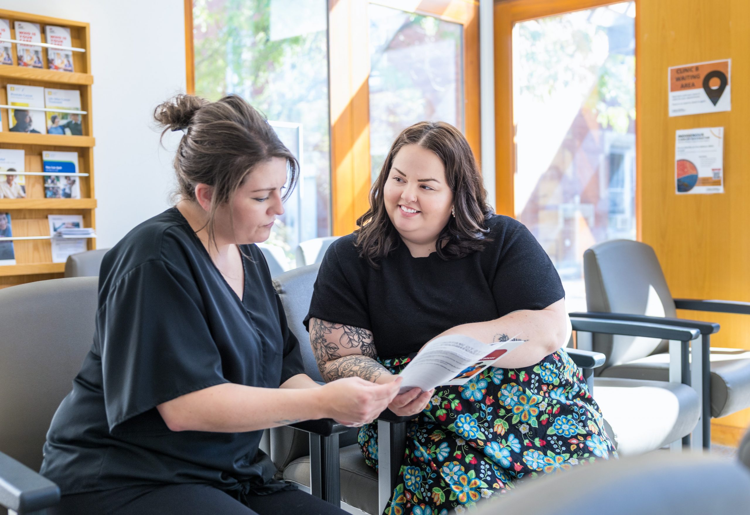 Two women sit in a waiting room, one holding and discussing a brochure with the other.