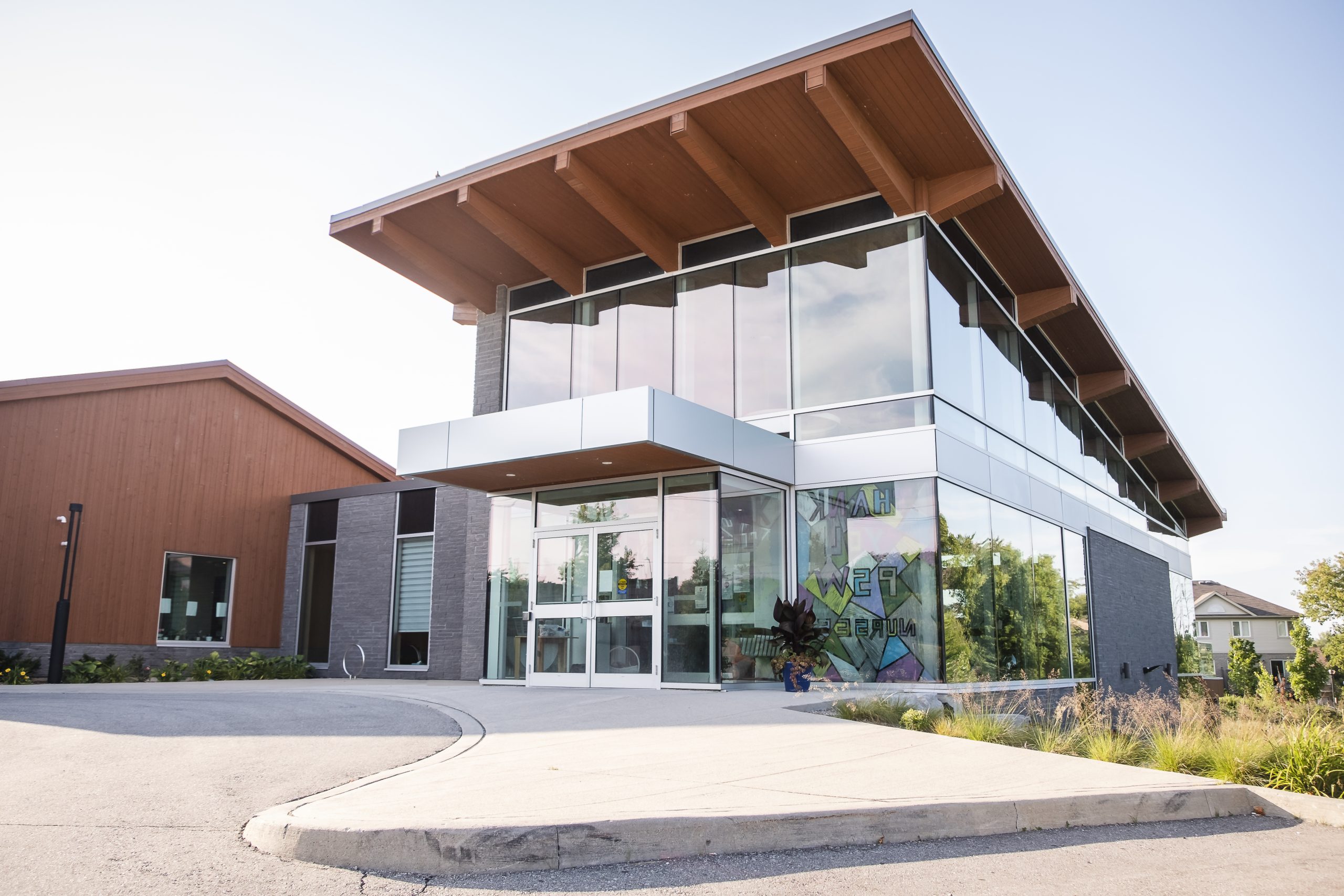 Modern building with large glass windows and a wooden overhang, set beside a parking lot on a sunny day.