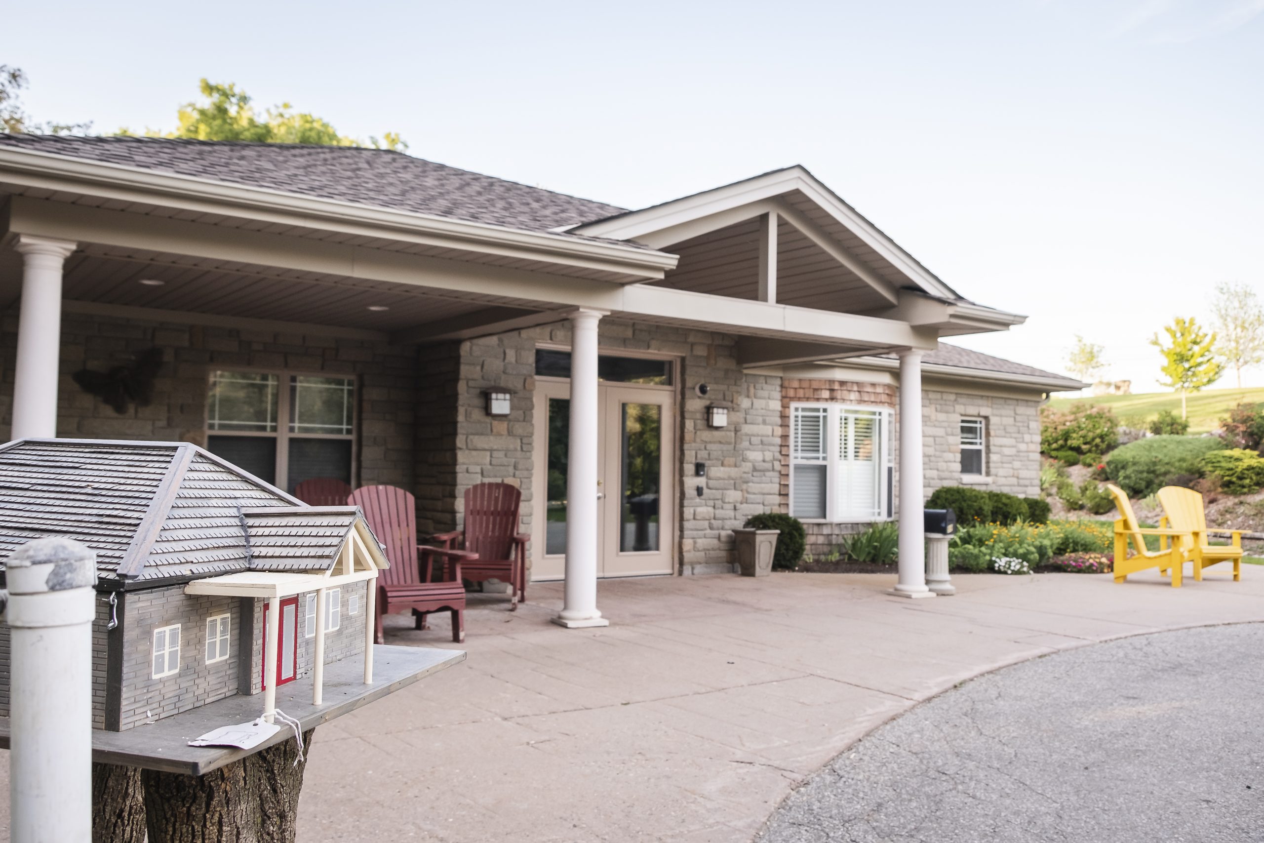A stone house with a front porch, red and yellow chairs, and a mailbox shaped like a house in the foreground.