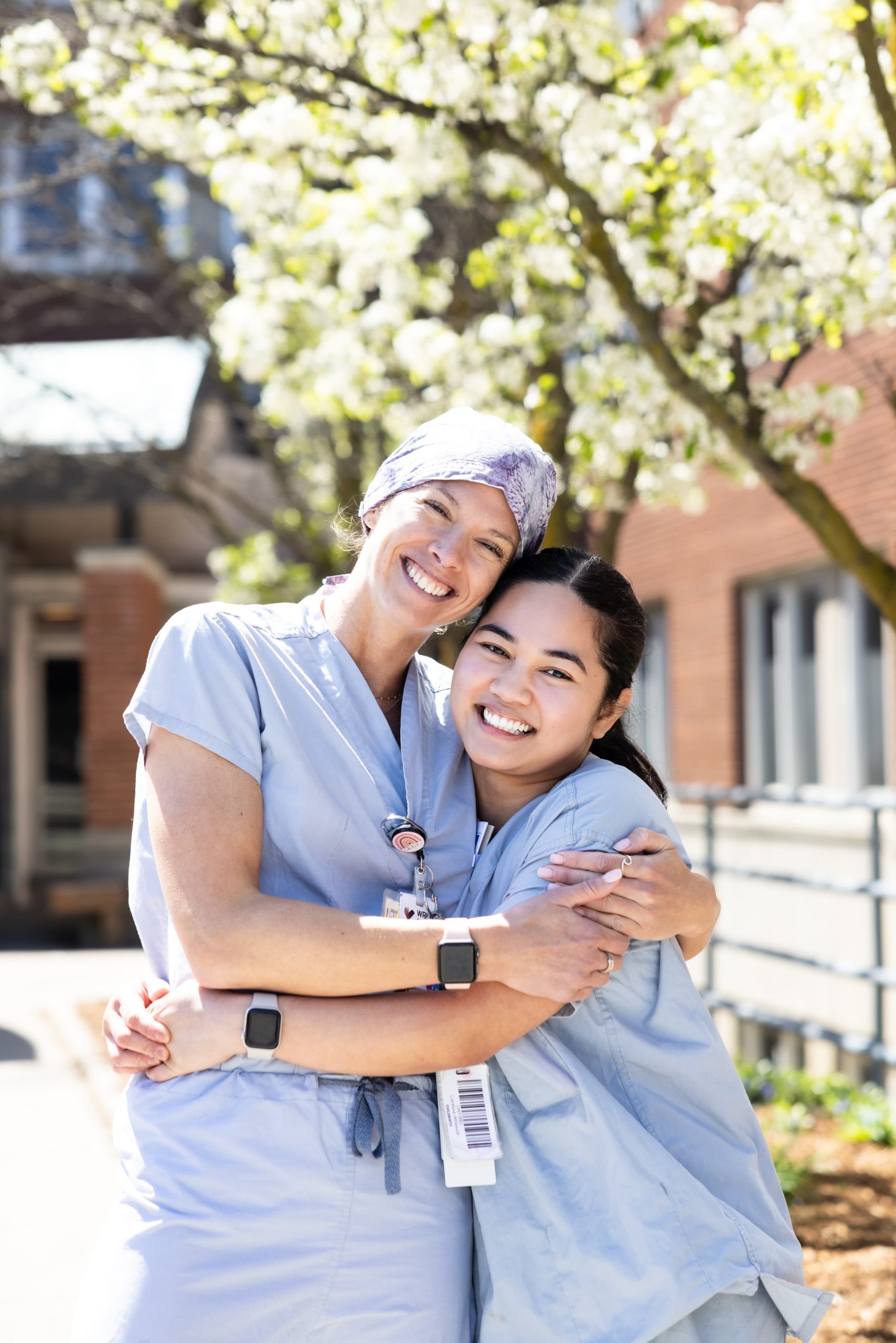 Two healthcare workers in scrubs smiling and hugging outdoors in front of a blossoming tree.