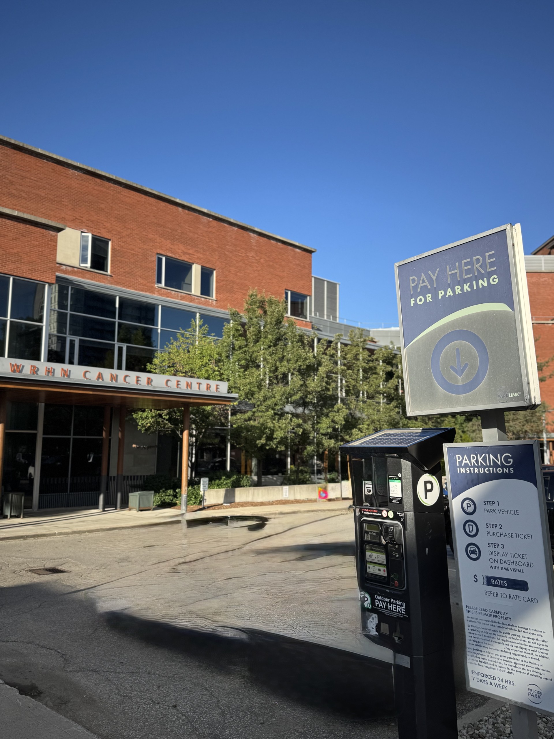 Parking payment machine in front of a brick building labeled "Cancer Centre" under a clear blue sky.