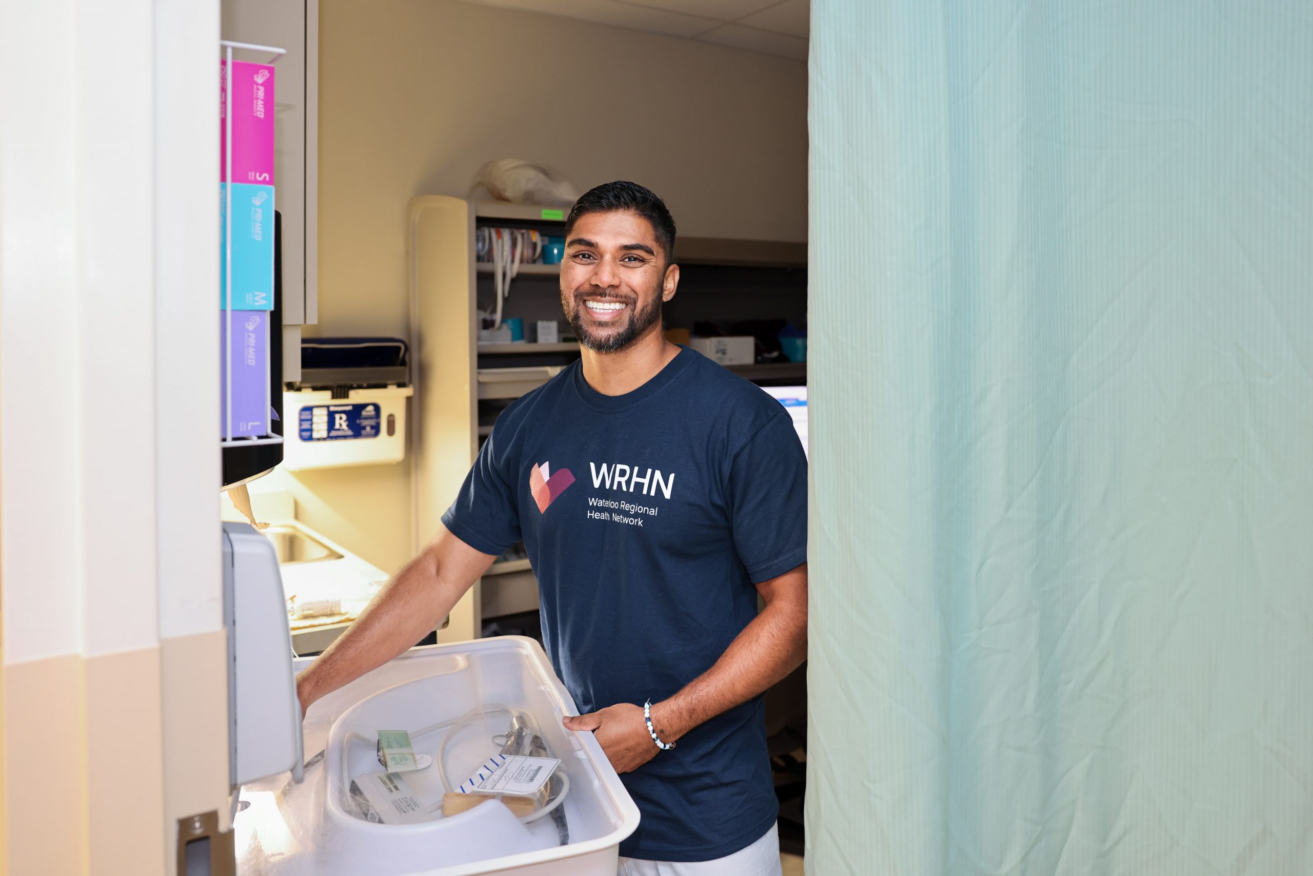 A man in a VRHN t-shirt smiles while standing next to medical supplies in a hospital room.