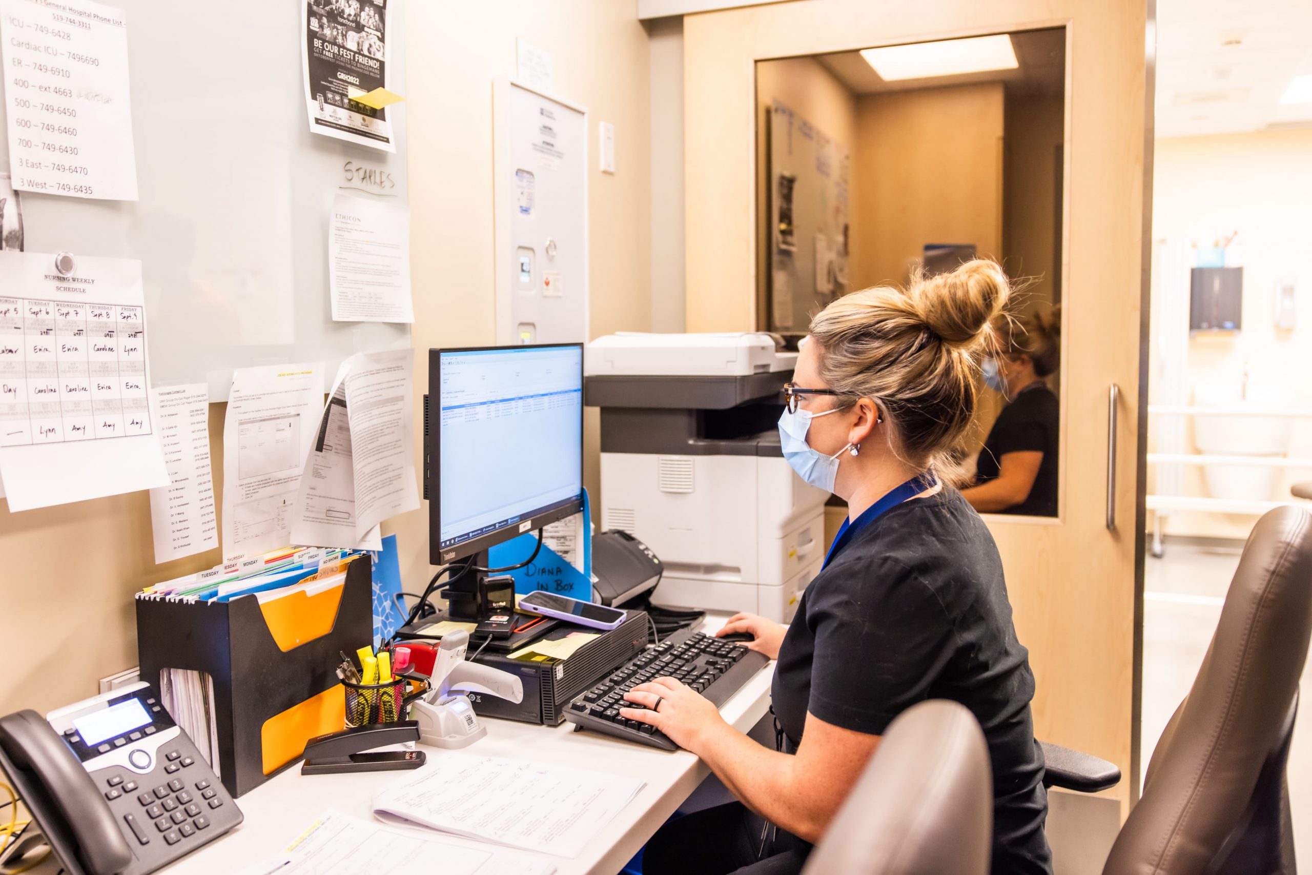 Woman wearing a mask works on a desktop computer at an office desk with papers, phone, and office supplies.