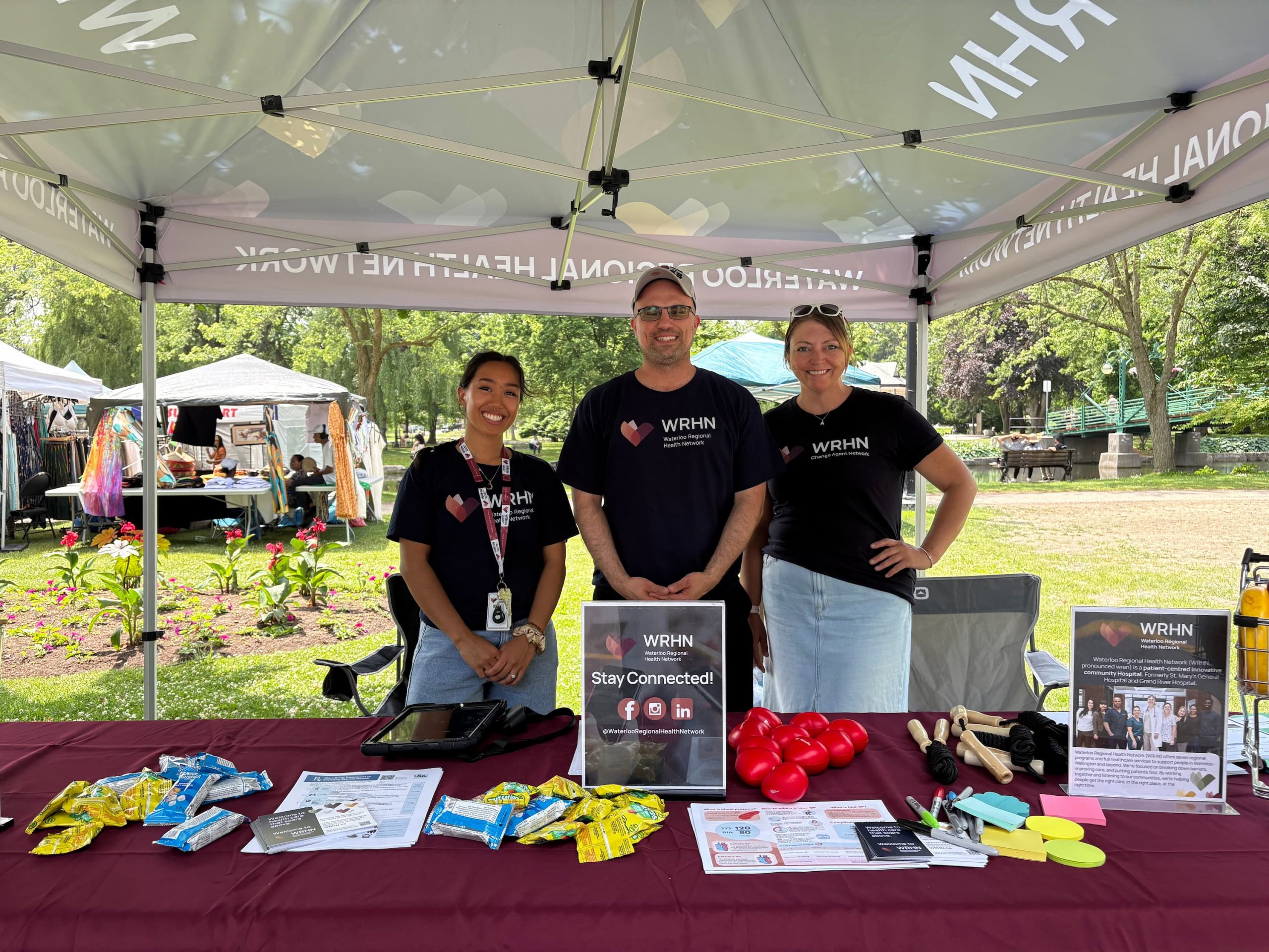 Three people stand behind a table with pamphlets and giveaway items at an outdoor informational booth under a canopy.