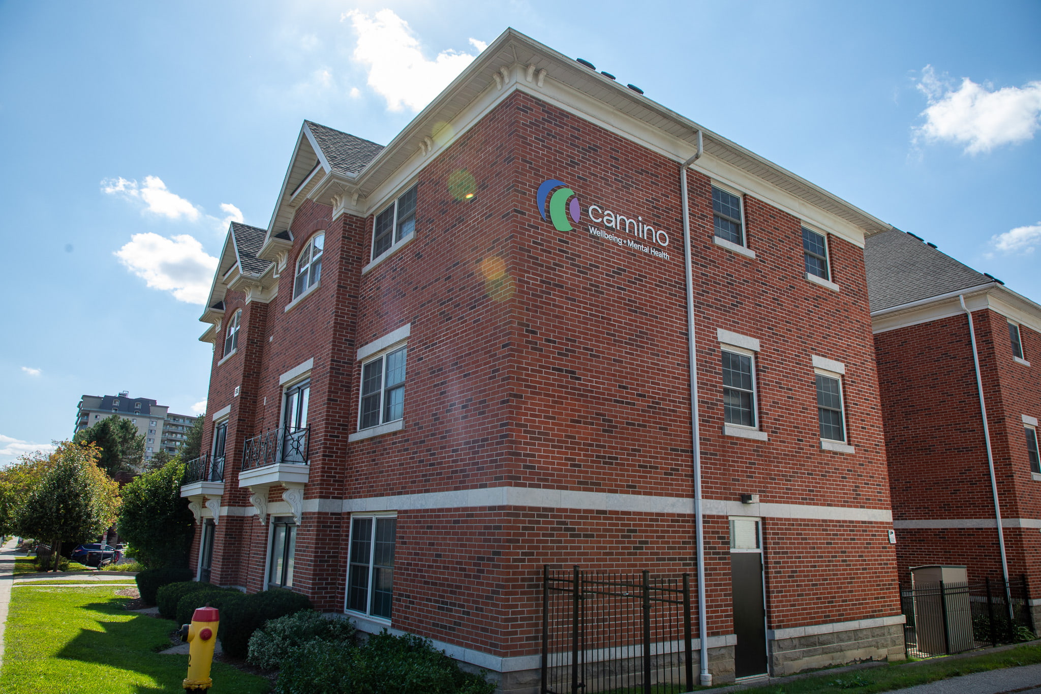 A three-story red brick building with a "Camino" sign on the side, set against a blue sky with scattered clouds.