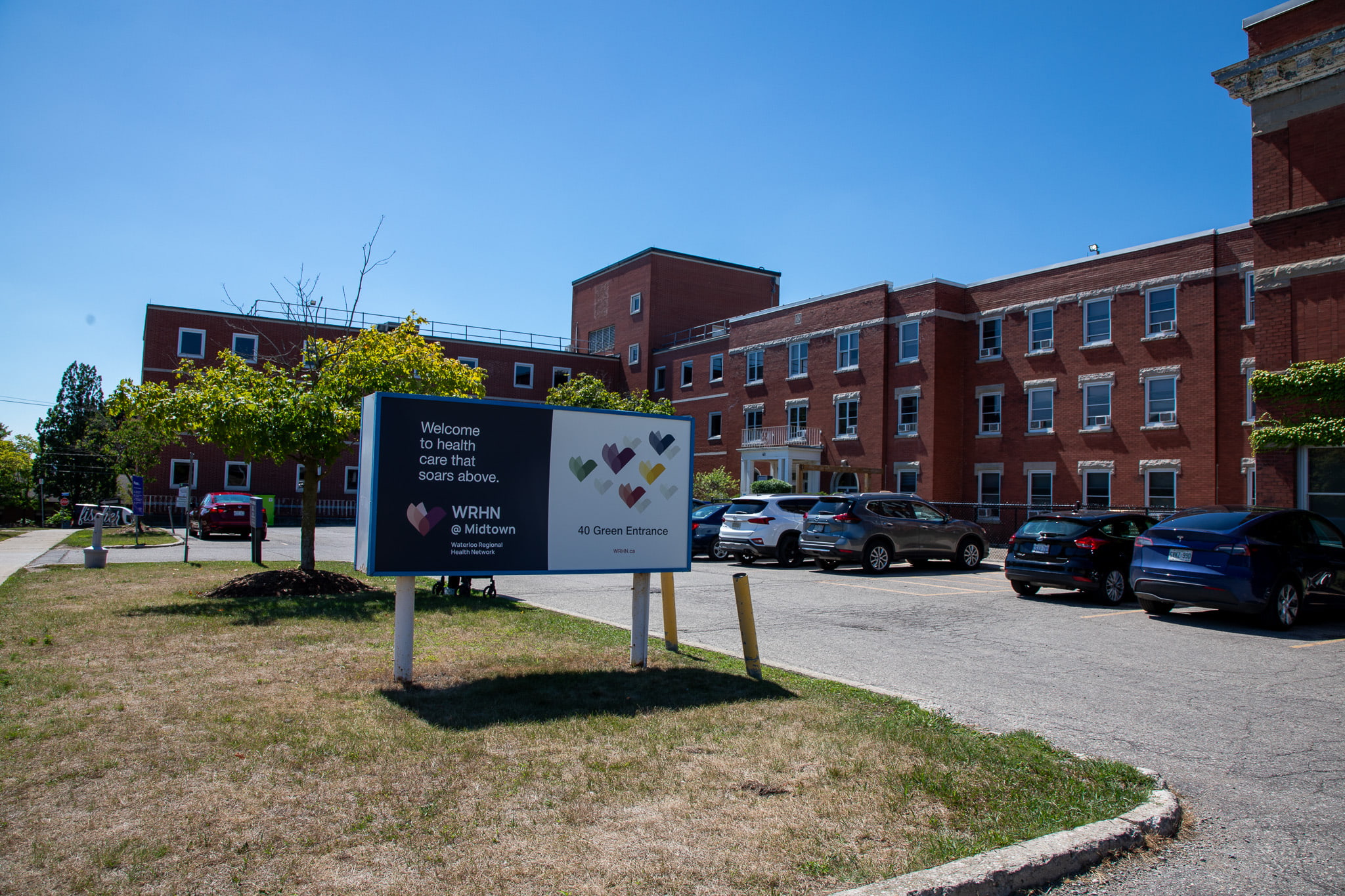 A brick healthcare facility with a parking lot and a sign reading "Welcome to health care that serves above" at the entrance.
