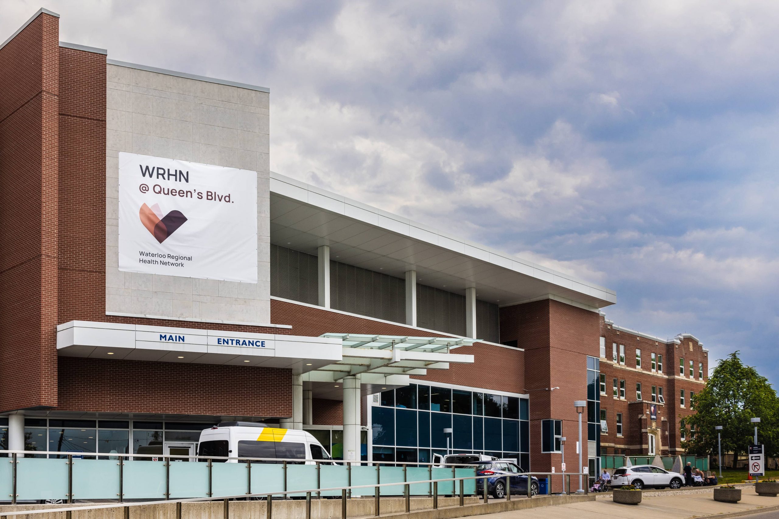 A modern hospital building with a sign reading "WRHN @ Queen’s Blvd." and vehicles parked outside the main entrance.