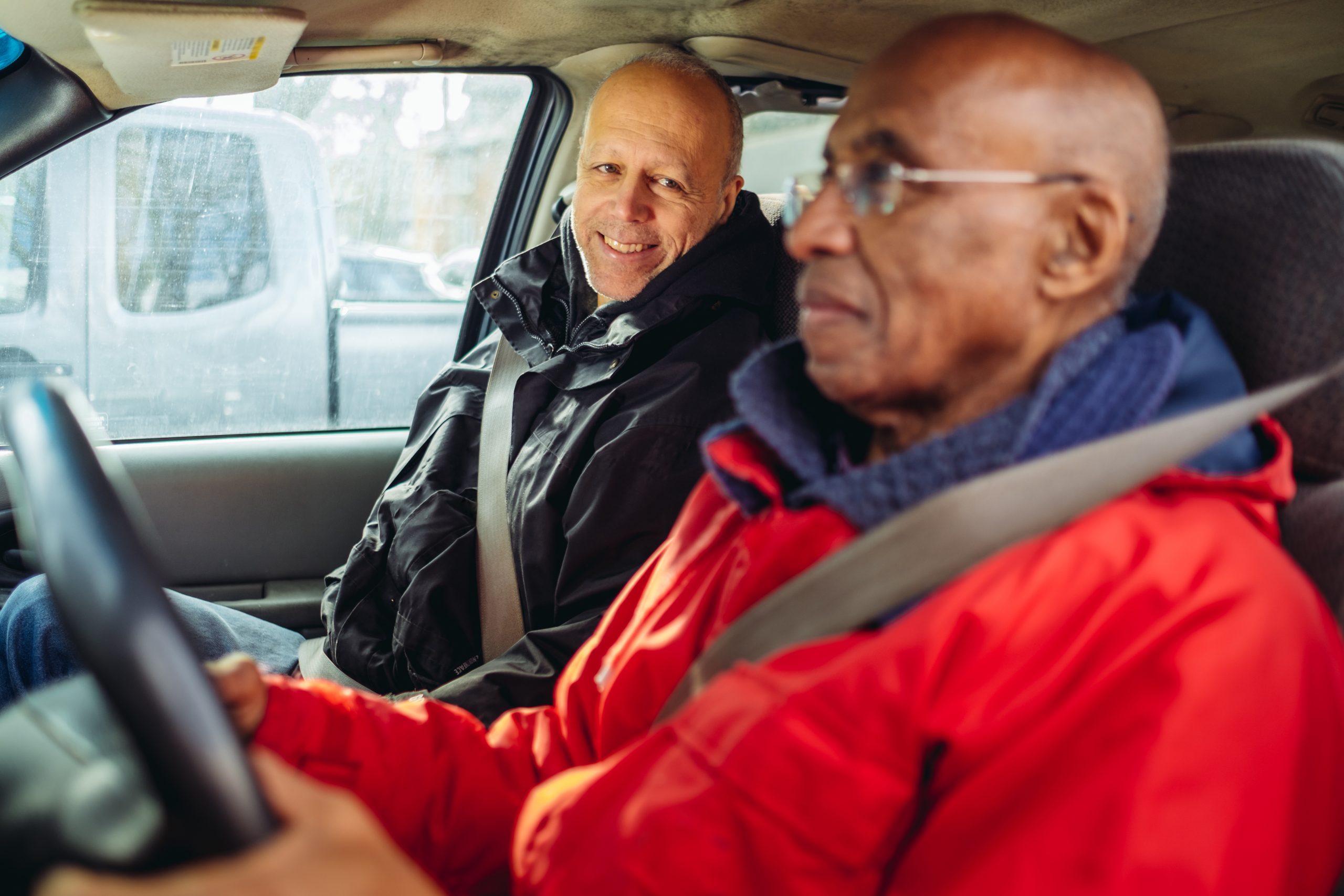 Two men wearing jackets and seat belts sit in a car, one driving and the other sitting in the passenger seat, both smiling.