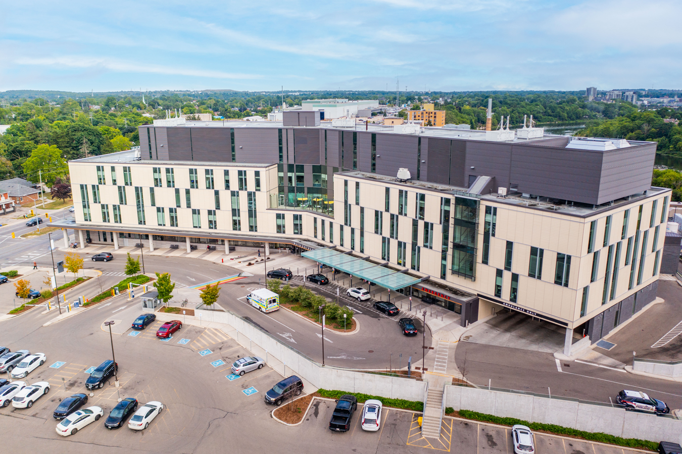 A large modern hospital building with parked cars in the lot, surrounded by greenery under a blue sky.