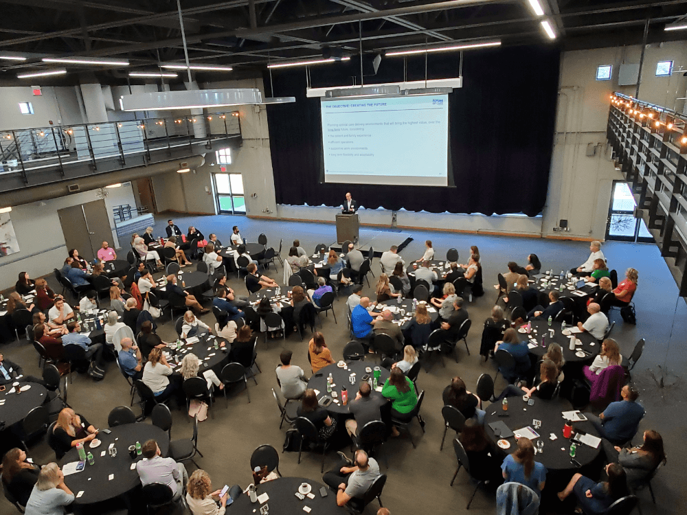 Large group of people seated at round tables listening to a speaker presenting slides in an event hall.