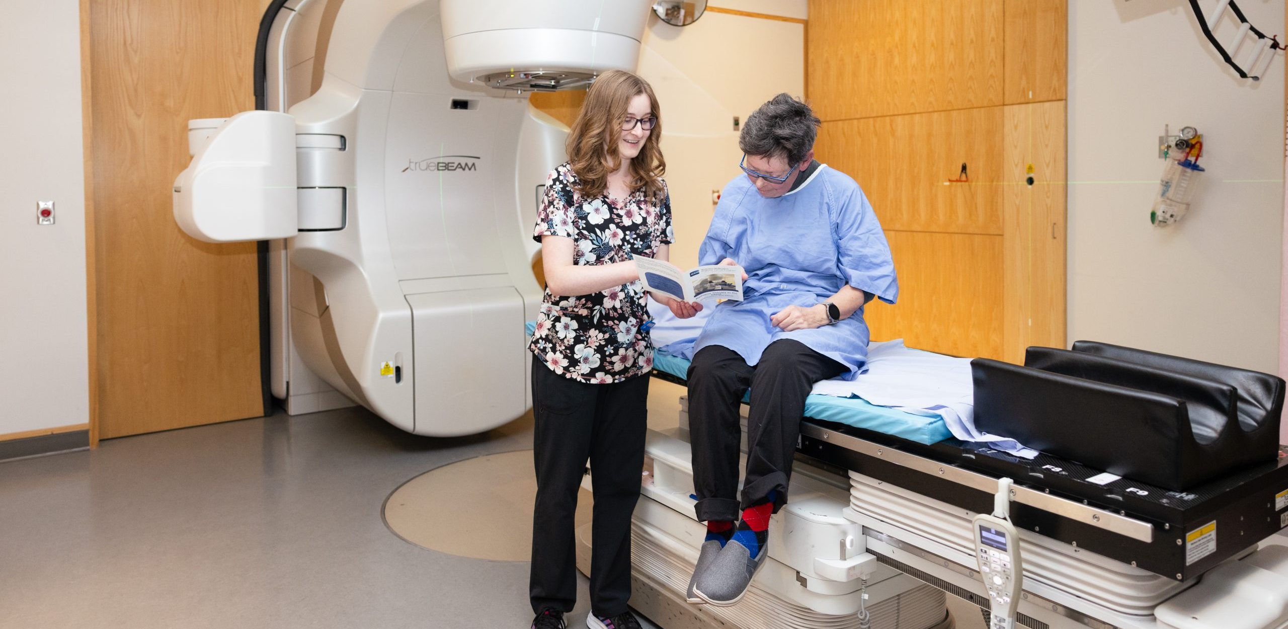 A healthcare worker discusses paperwork with a patient sitting on a treatment table in a medical imaging room.