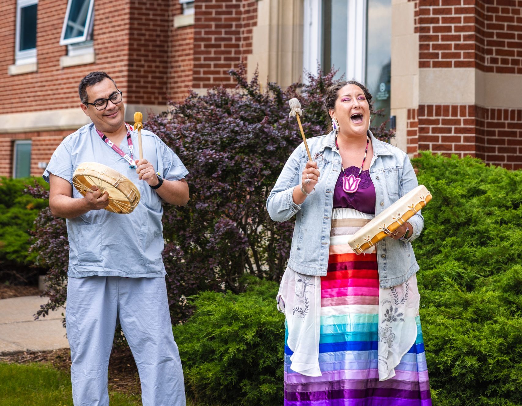 Two people stand outside a brick building, each playing a hand drum and smiling, with a WRHN sign above the entrance.