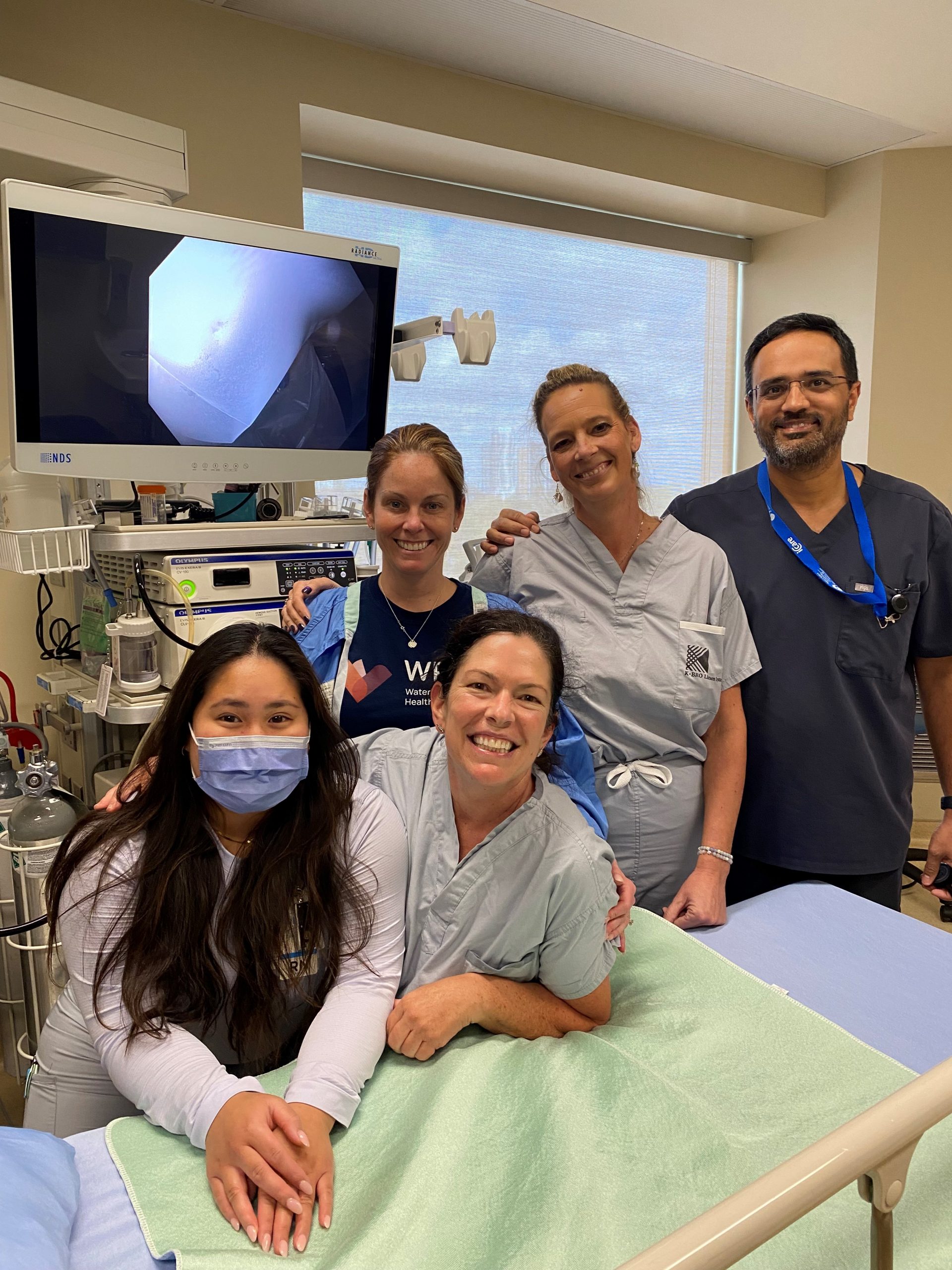 Five medical professionals pose and smile in a hospital room, with medical equipment and a monitor in the background.