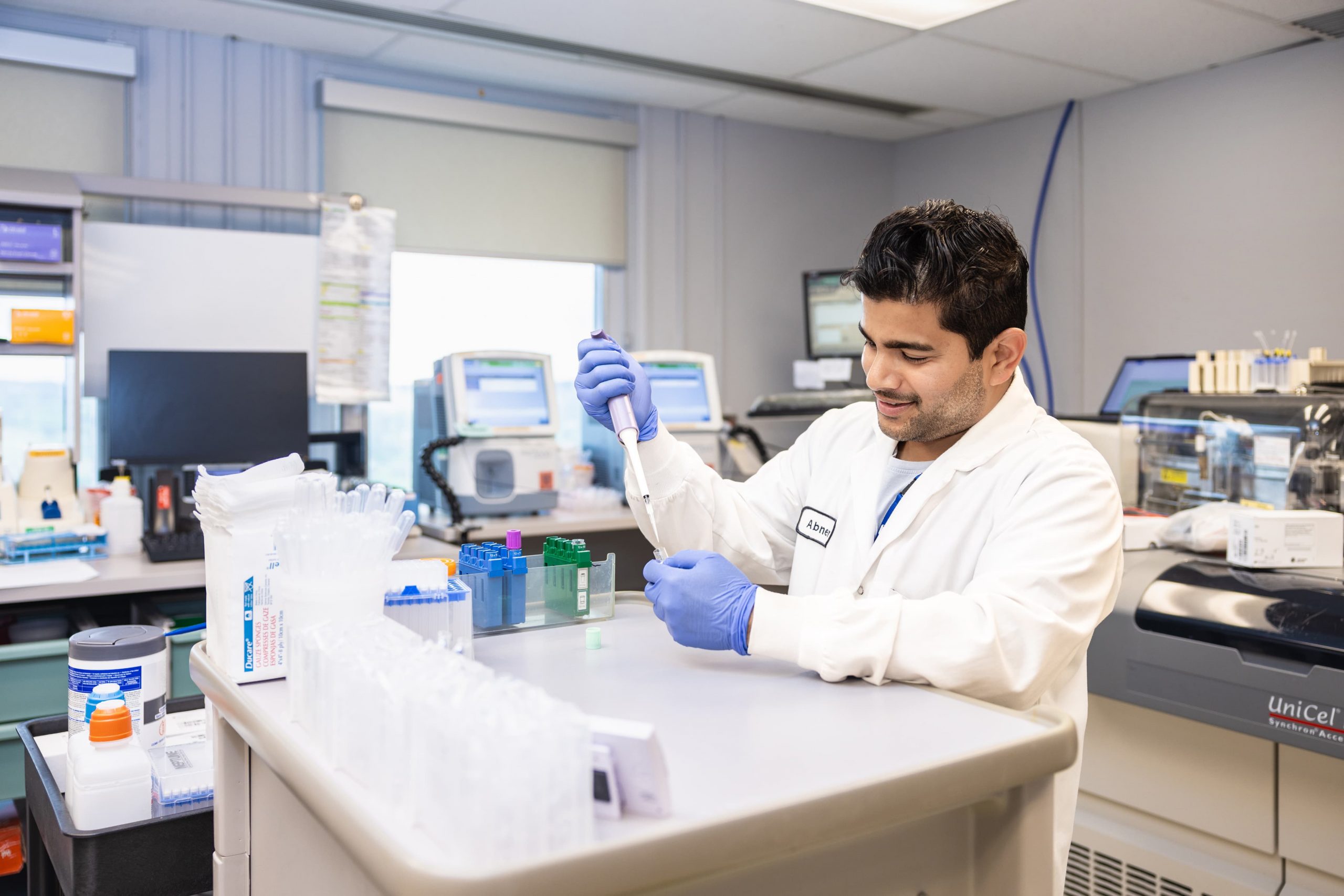 A scientist in a lab coat and gloves uses a pipette to transfer liquid into a test tube in a laboratory setting.