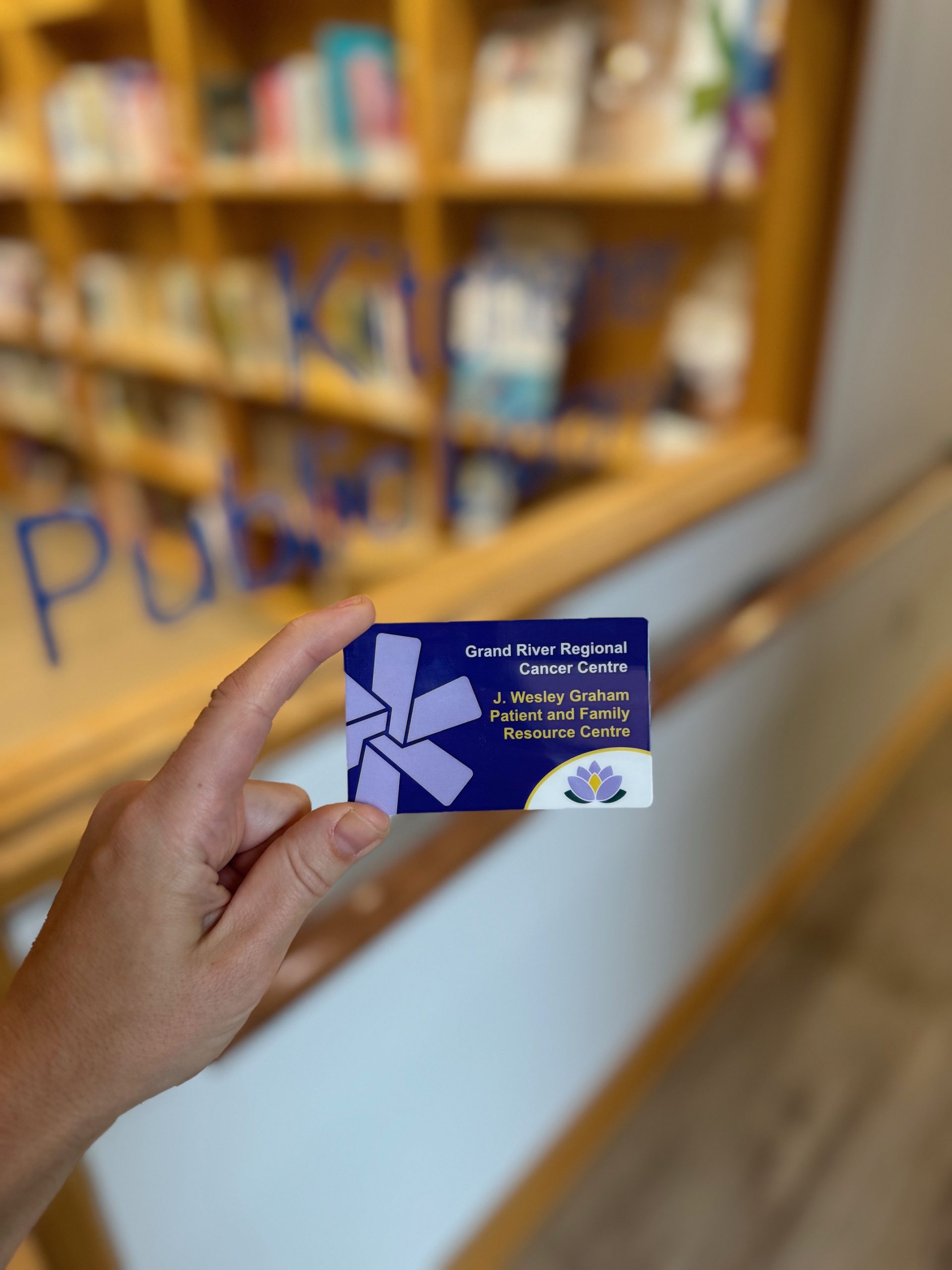 A hand holds a Grand River Regional Cancer Centre card in front of a blurred library or bookstore background.