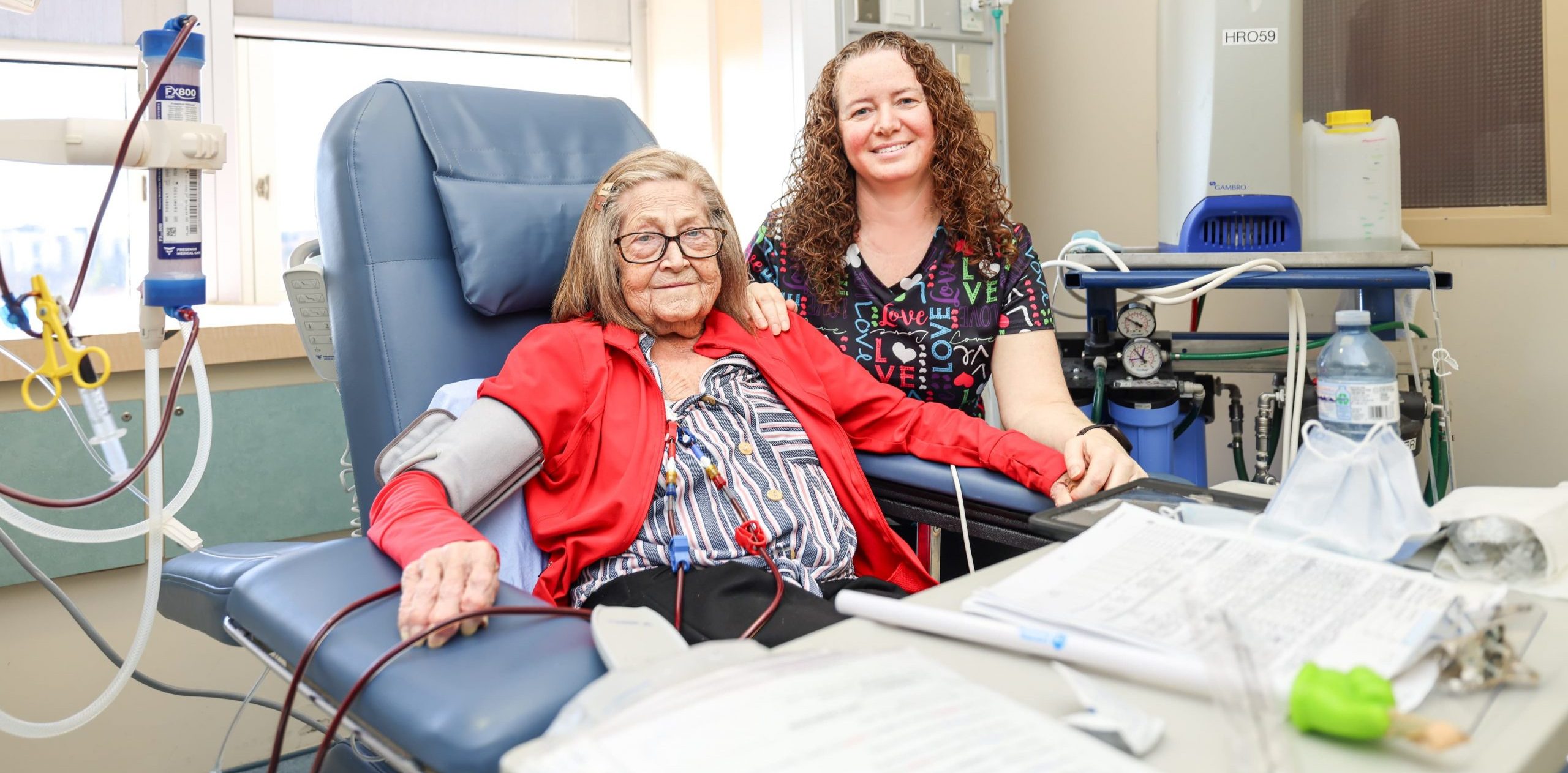 A patient receives medical treatment in a clinic, seated beside a smiling healthcare worker.