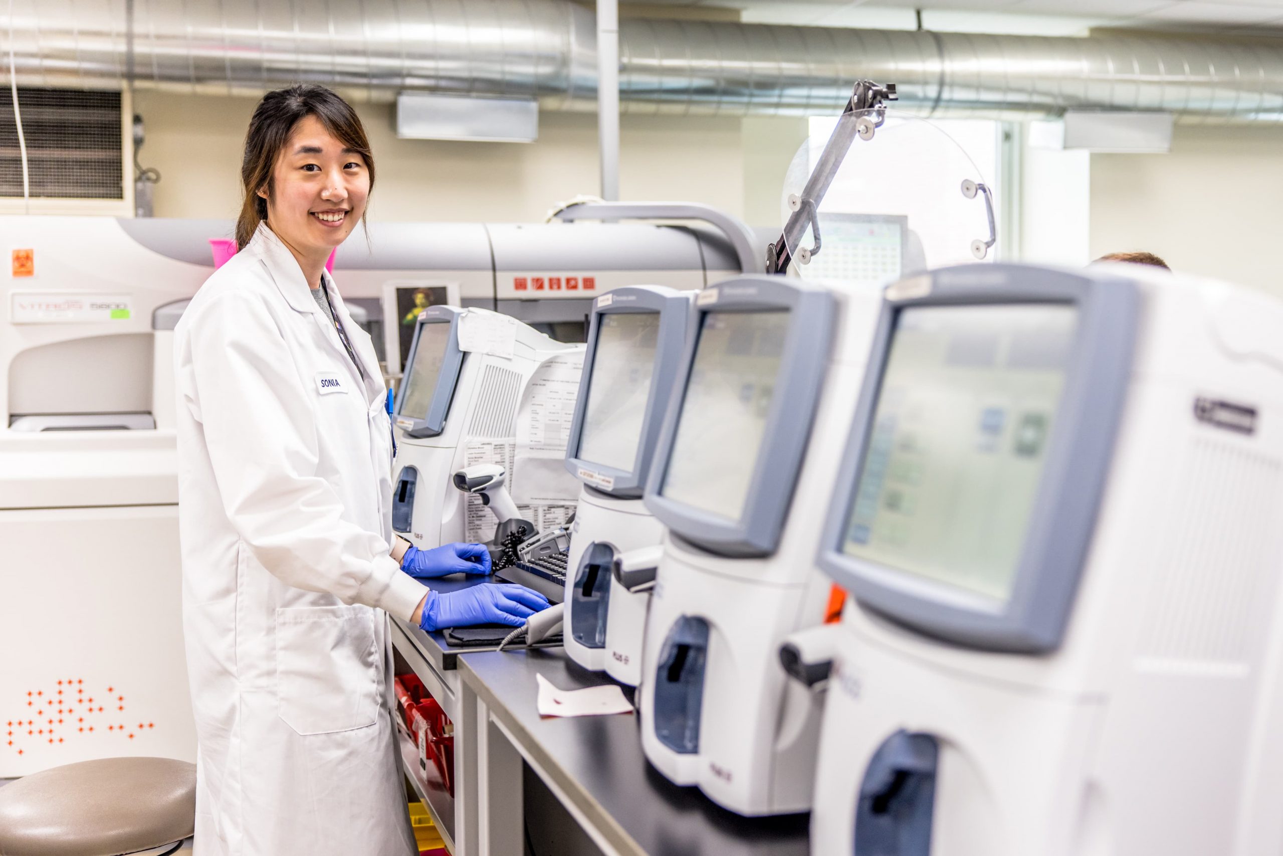 A lab technician in a white coat and gloves operates medical diagnostic machines in a laboratory setting.