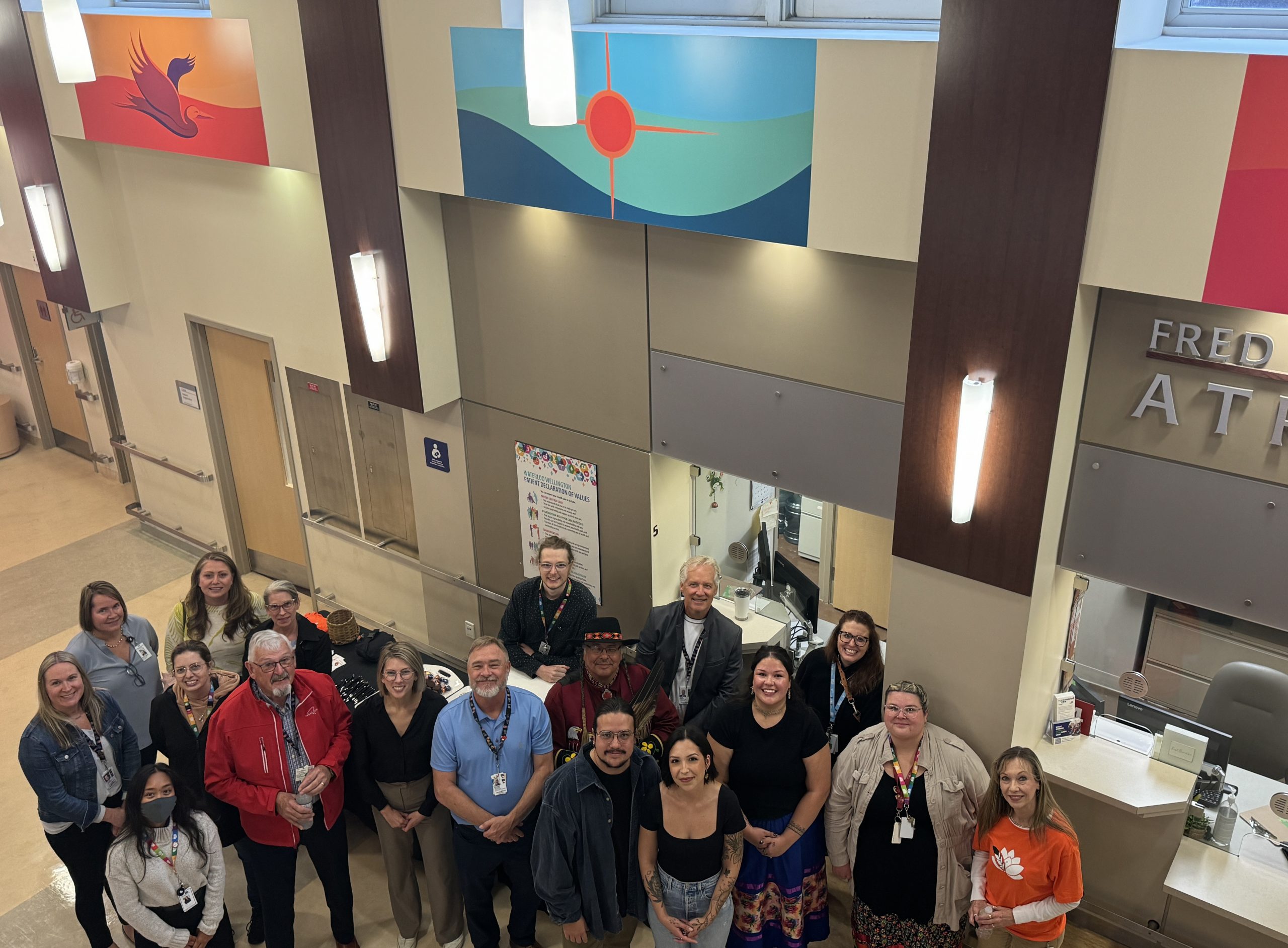 A group of adults stands together in a lobby area, some smiling for the camera, beneath colorful artwork on the walls.