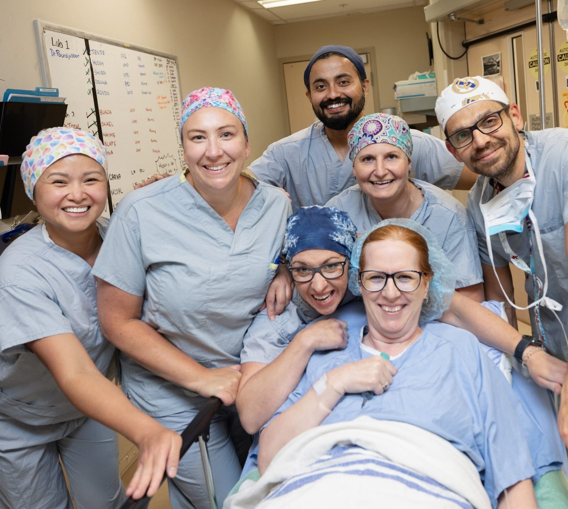 A group of healthcare professionals smiles around a patient in a hospital setting.